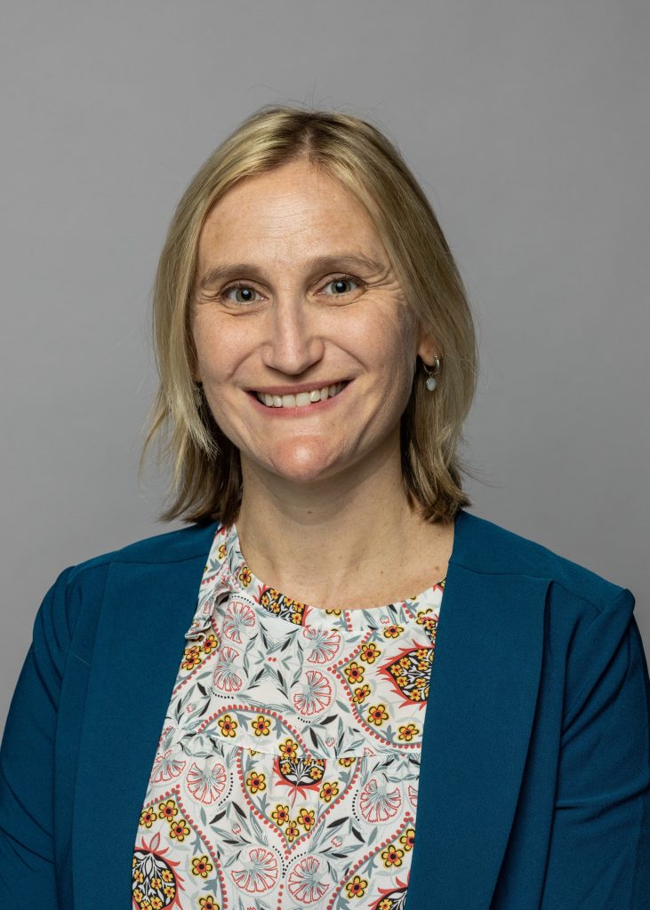 Julia Stensby, a smiling woman with should length blonde hair, wearing a blue blazer and a shirt with a multicolored pattern.