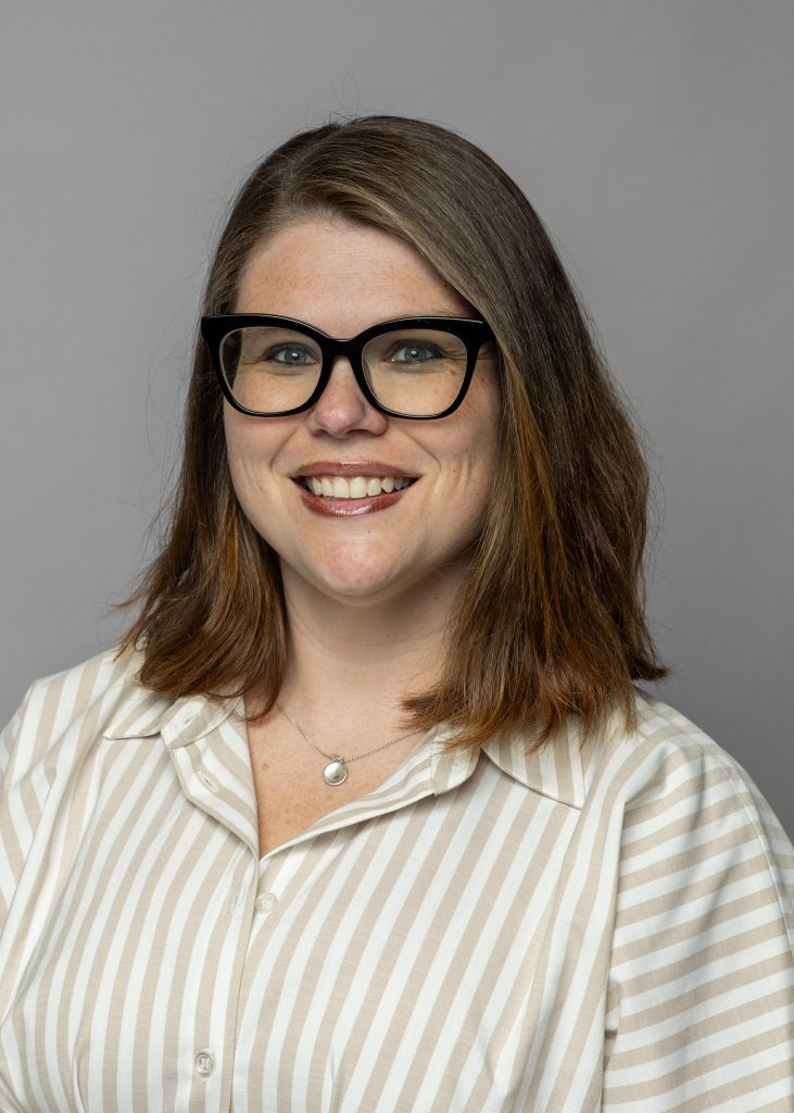 Katryna Sardis, a smiling woman with shoulder length brown hair, wearing a tan and white striped dress and glasses.
