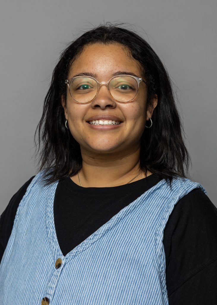 Vanessa Davis, a smiling woman with shoulder length wavy black hair, wearing glasses, a black shirt, and a denim dress.